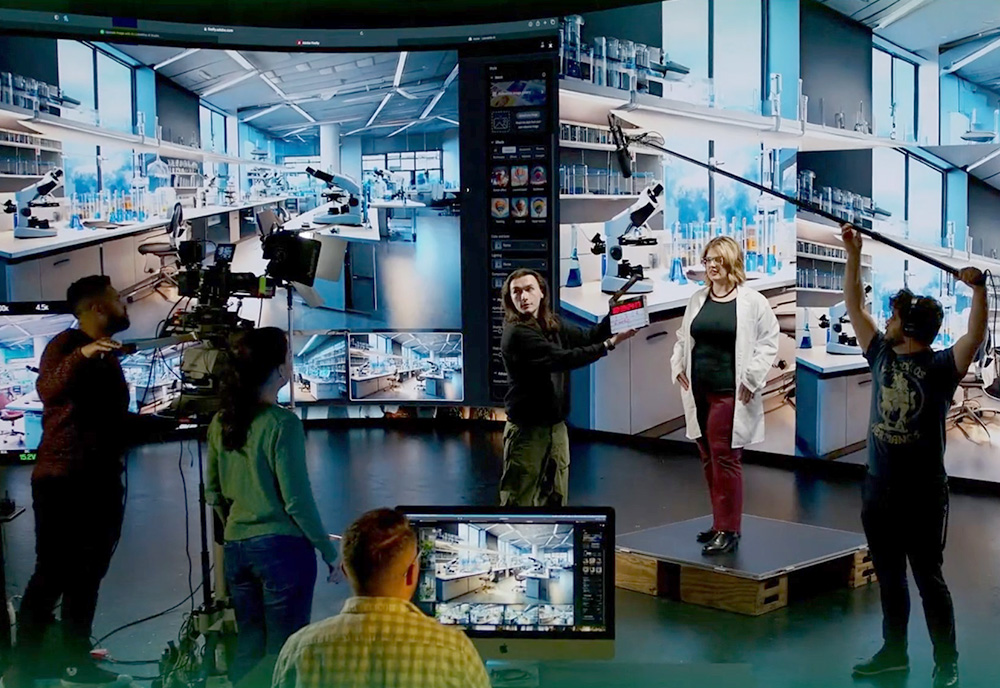 A group of students in a televison production studio record a woman in front of a digital screen